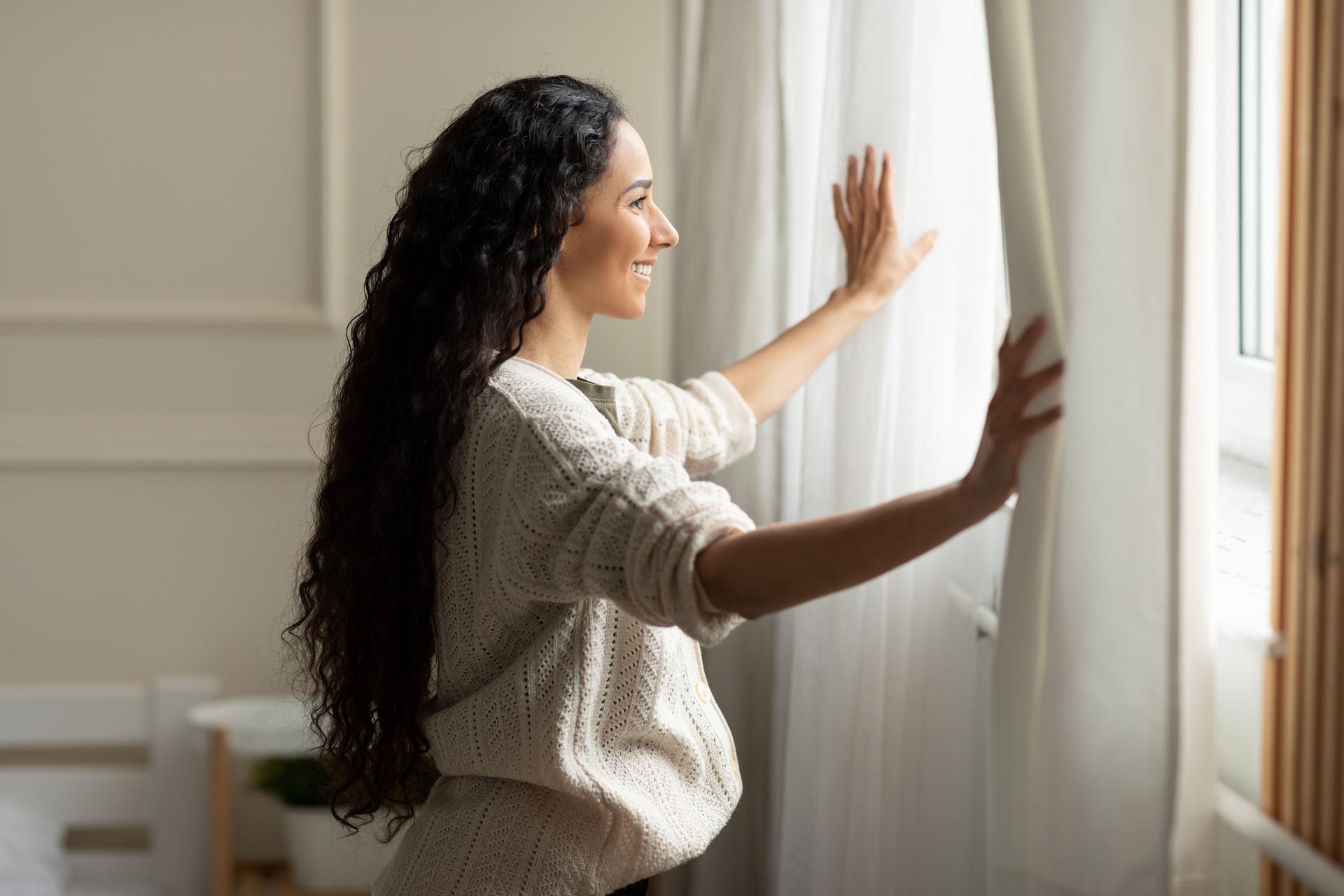 Smiling woman opening window curtains at home