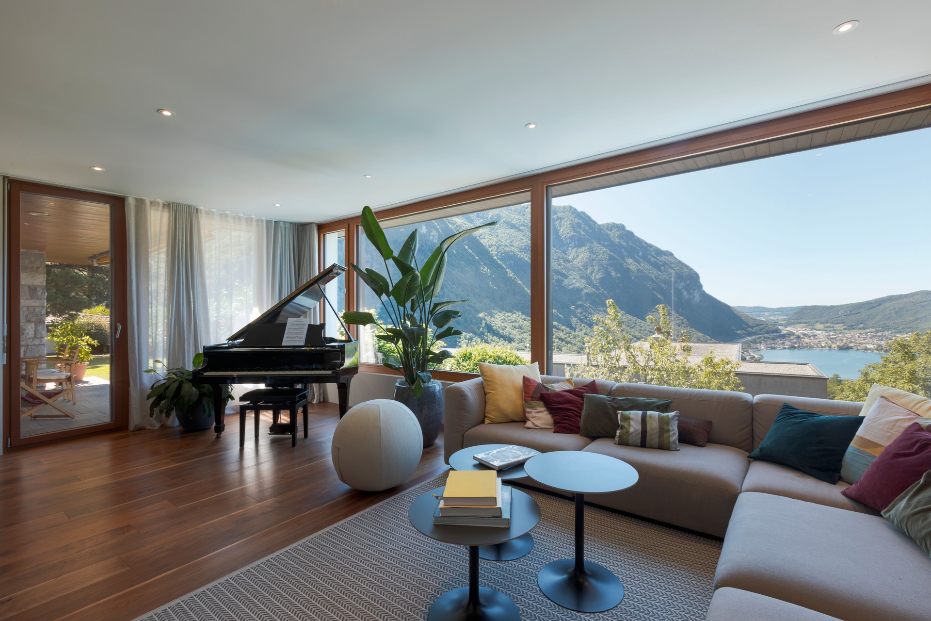 Living room with large light-colored sofa and lots of cushions and a black piano. Large window overlooking the valley
